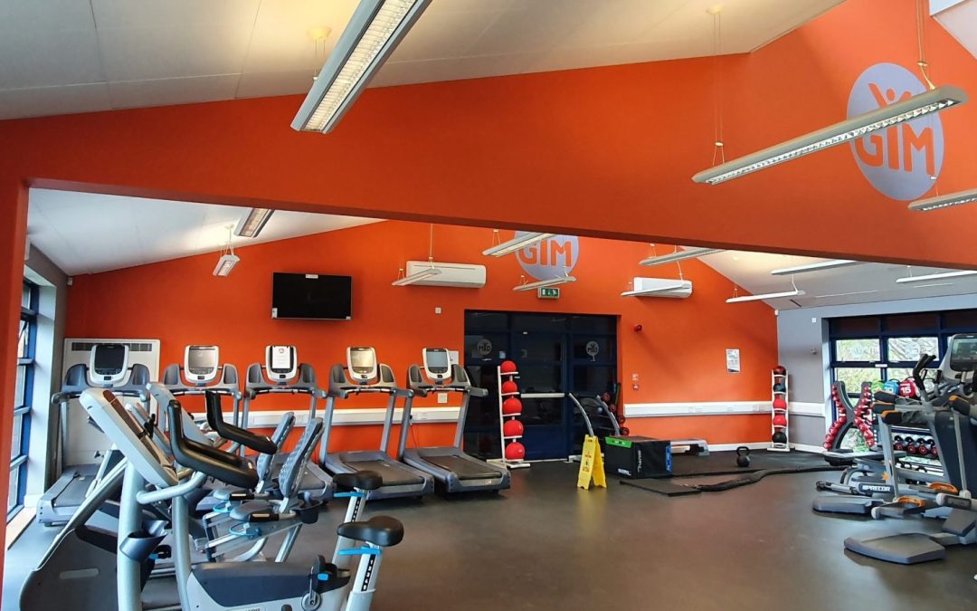 Bright gym interior with orange branded GIM walls featuring rows of treadmills, cardio machines, and a wall-mounted TV