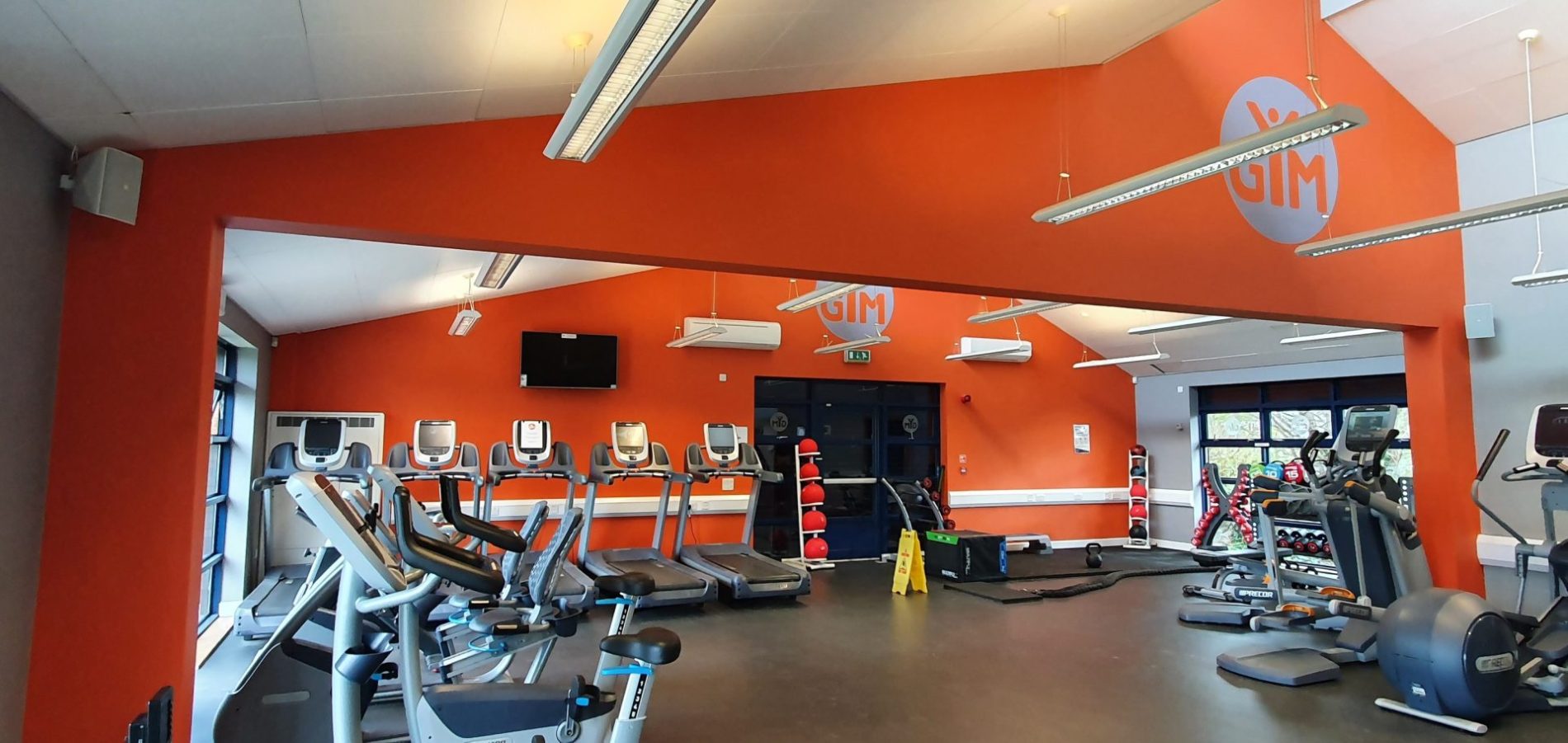 Bright gym interior with orange branded GIM walls featuring rows of treadmills, cardio machines, and a wall-mounted TV
