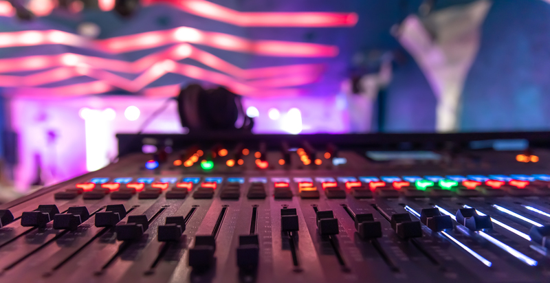 Close-up of a professional audio mixing console with colorful faders and LED indicators, with blurred pink stage lighting and a performer in the background