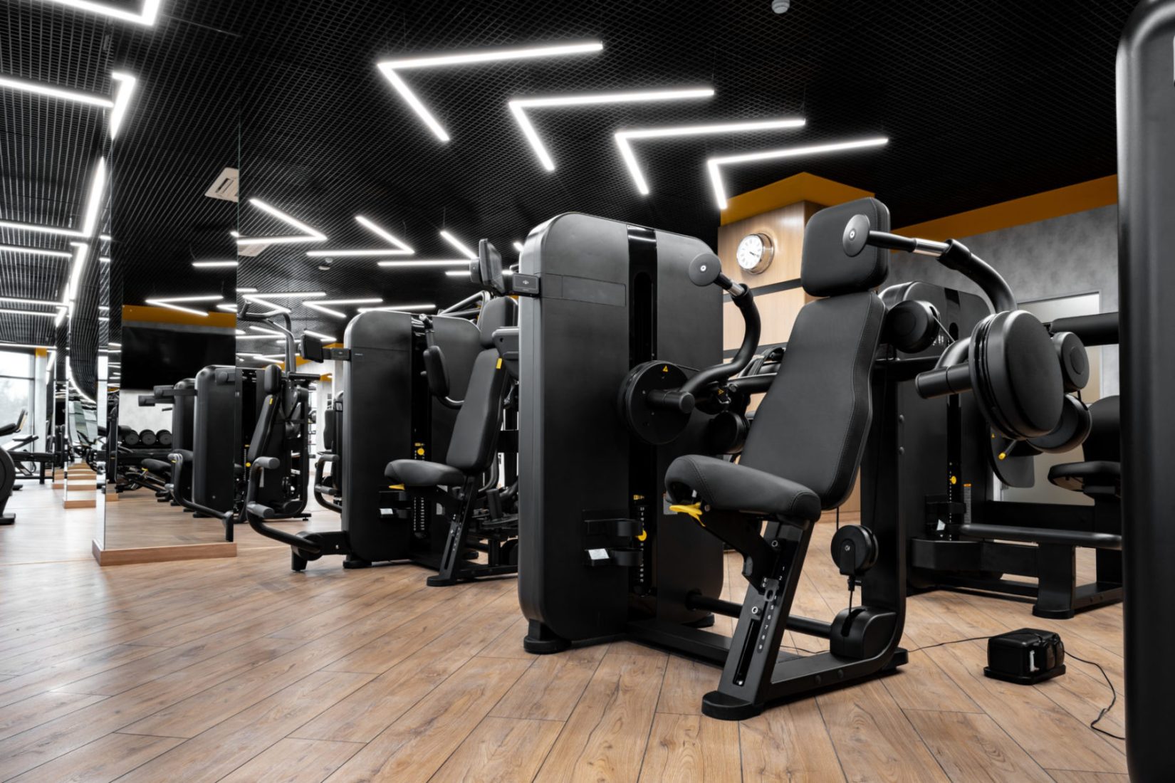 Modern gym interior with new fitness equipment Modern gym interior with rows of black and gold strength training machines on hardwood floors under geometric LED ceiling lights