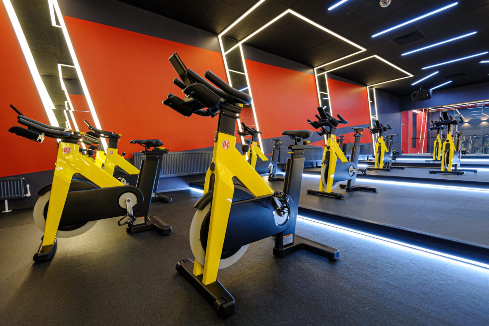Spin studio with yellow and black stationary bikes arranged in rows against a bold red accent wall with geometric LED lighting
